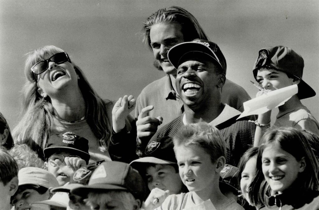 A photograph of Mike "Pinball" Clemons of the Toronto Argonauts with children and administrators from the Allenby Daycare Centre. The Toronto Star article that featured this photographed had the headline "Popular pinball: Argos standout Mike Clemons makes a bunch of the new friends from the Allenby Daycare Centre before yesterday's practice session" and was published September 4, 1992. Image by Rick Eglinton and sourced from the Toronto Public Library.